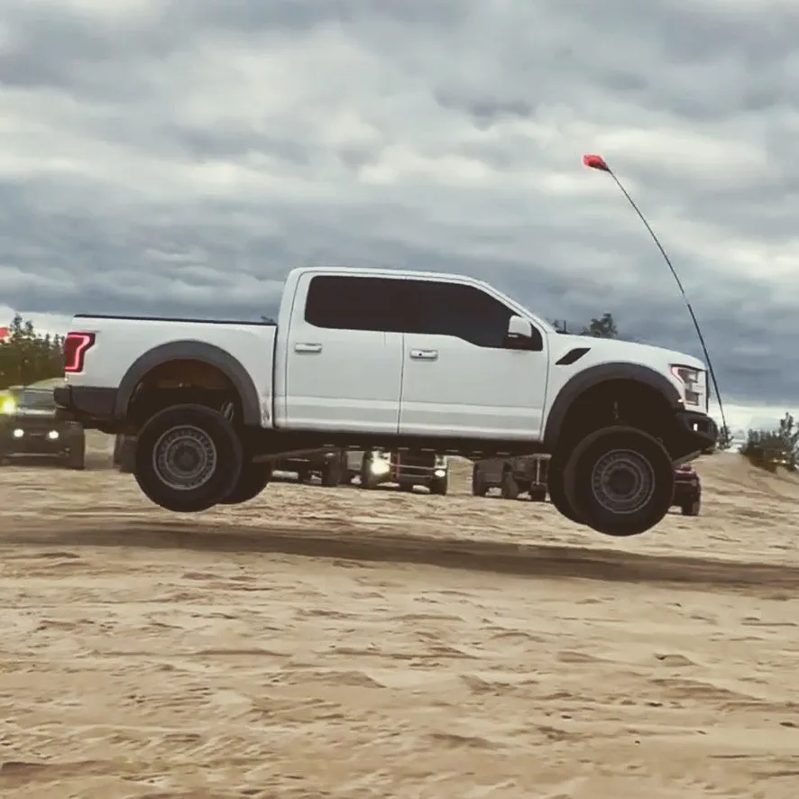 A white Ford Raptor airborne over a sand dune under a grey sky